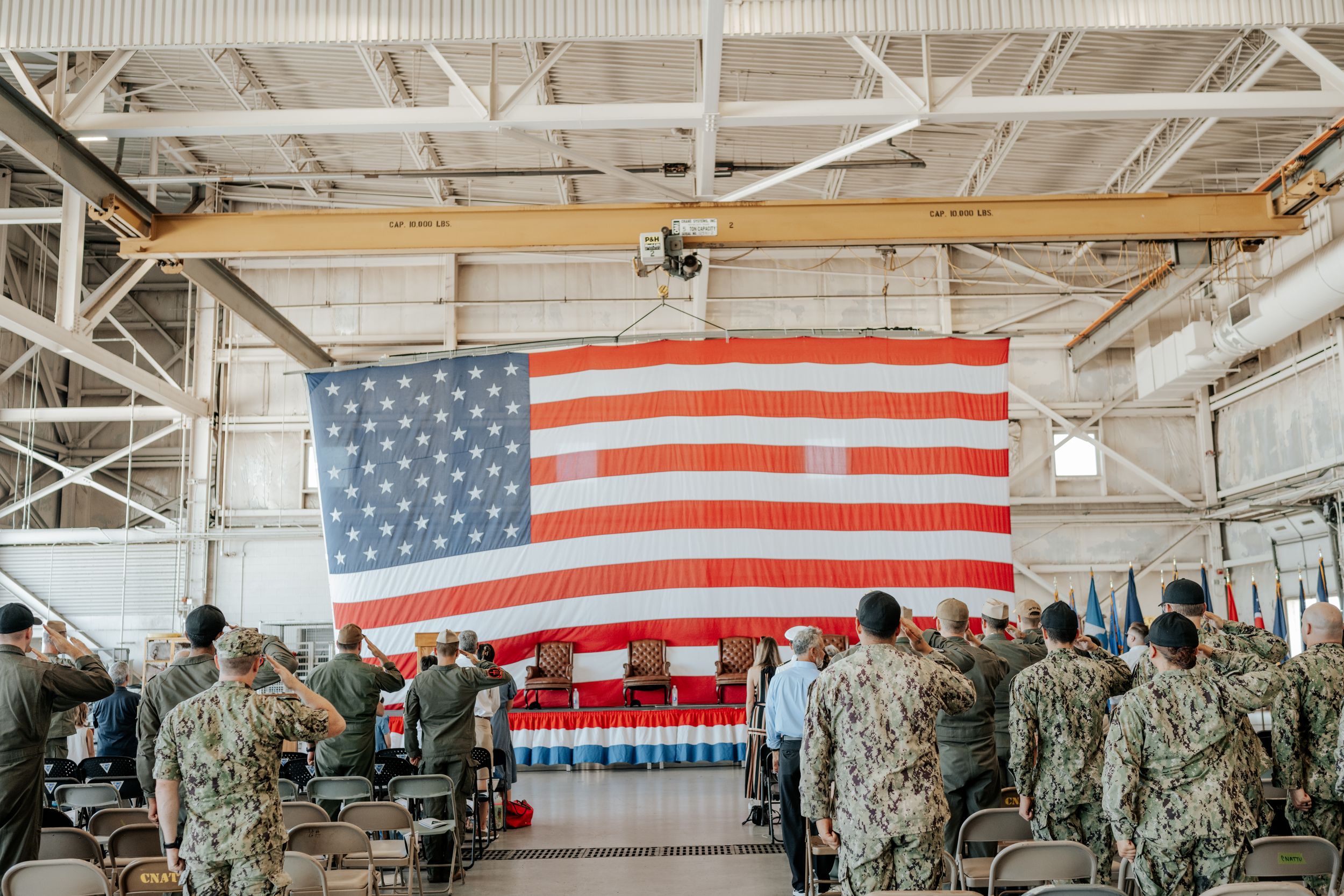 Military service members stand in formation facing a large American flag displayed inside an industrial building.