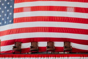 Four wooden chairs arranged in a row against a large hanging American flag background creating a patriotic display.