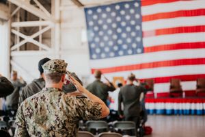 Military service members salute during a ceremony in front of a large American flag display indoors.