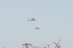 Two military helicopters flying in a grey sky above a construction site with visible cranes in the background.