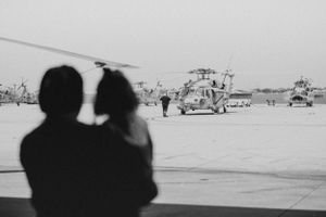 A silhouetted couple watches military helicopters on an airfield tarmac in black and white.