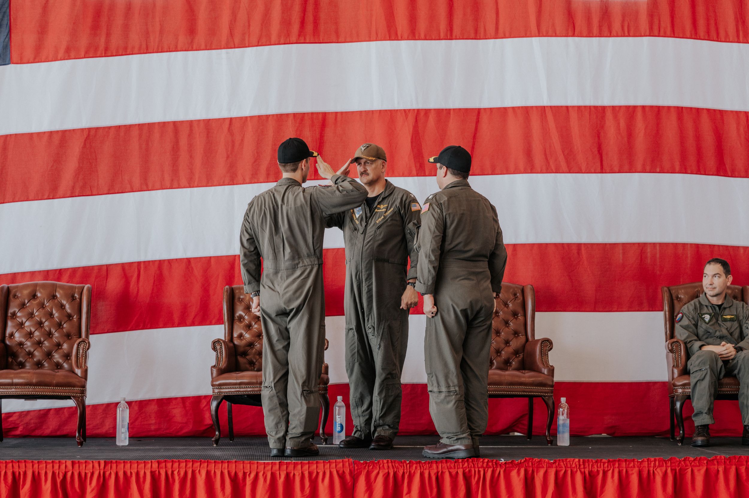 Military service members stand on a stage with a large red and white striped American flag backdrop during a ceremony.