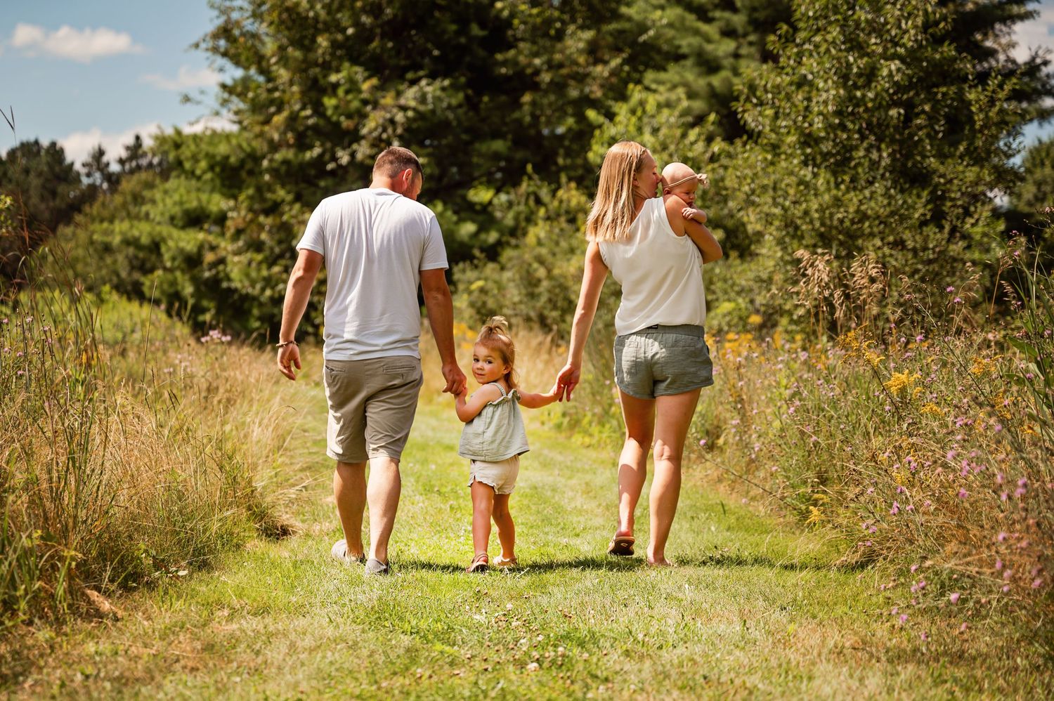 Family walking hand in hand on a sunny summer path through tall grass.