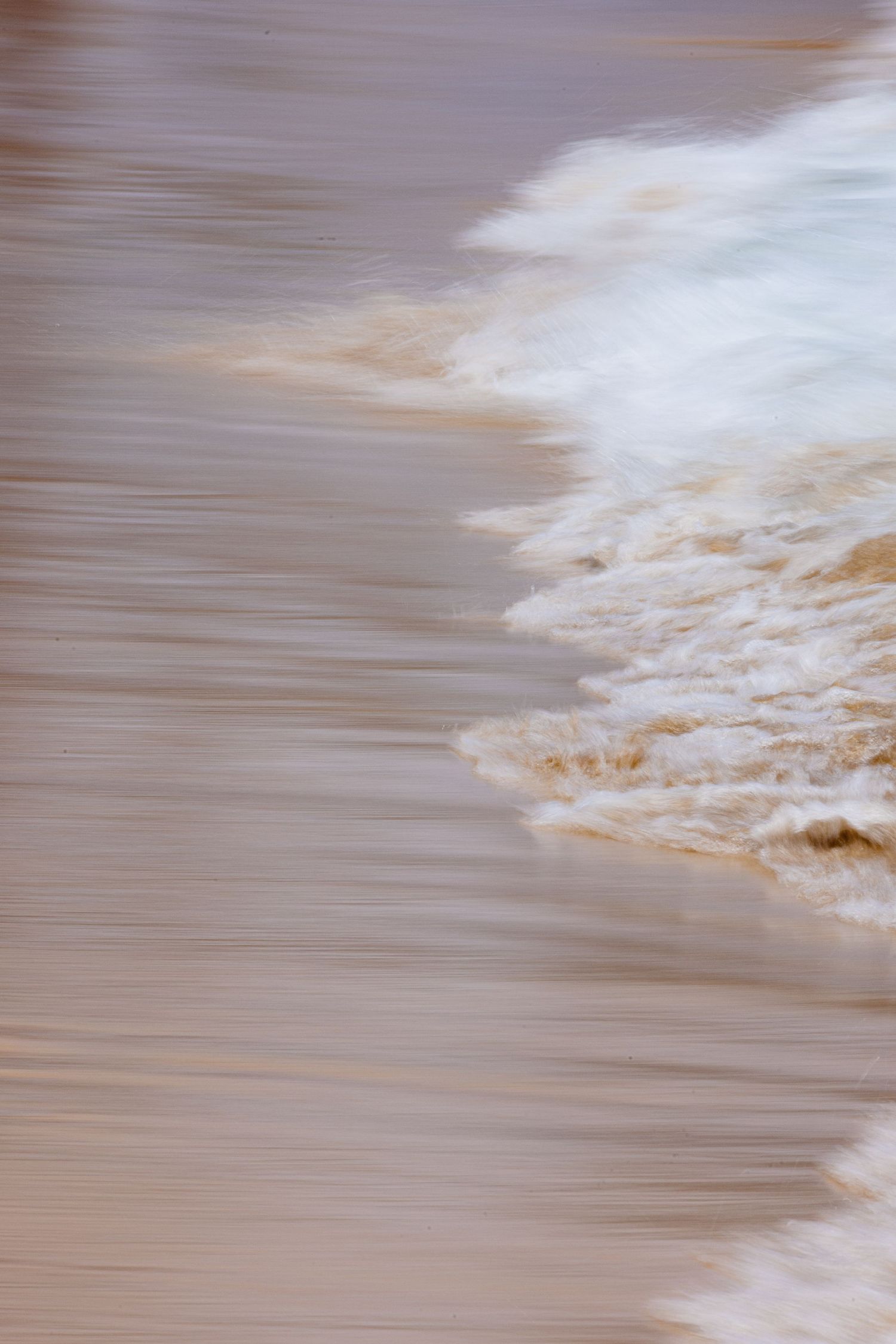 Blurred motion of sandy ocean waves washing ashore, creating an abstract pattern of white foam and brown water.