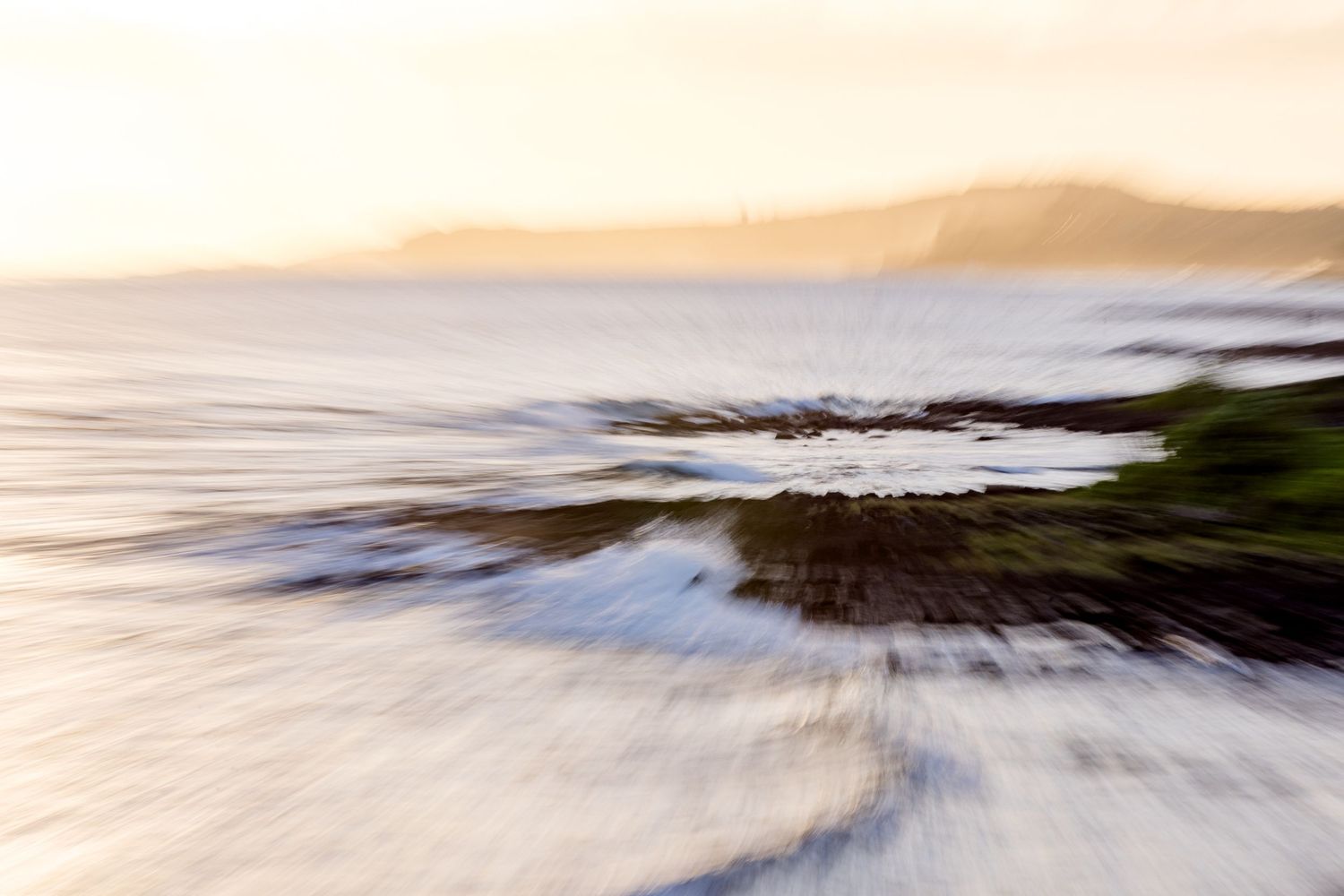 Blurred motion shot of ocean waves crashing on rocky shore at golden sunset with misty mountains in background.