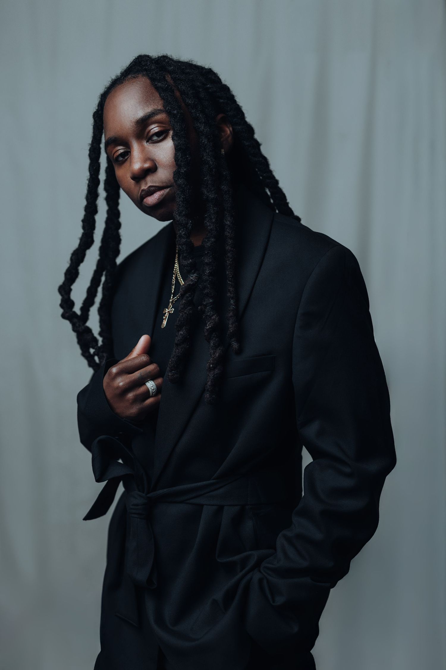 Person wearing black puffy jacket and cap poses against grey background in studio setting.