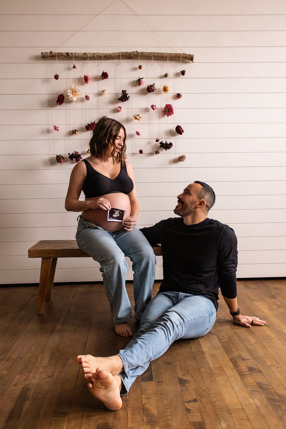 Expectant couple sits together on wooden bench with dried floral wall decor in rustic indoor setting.