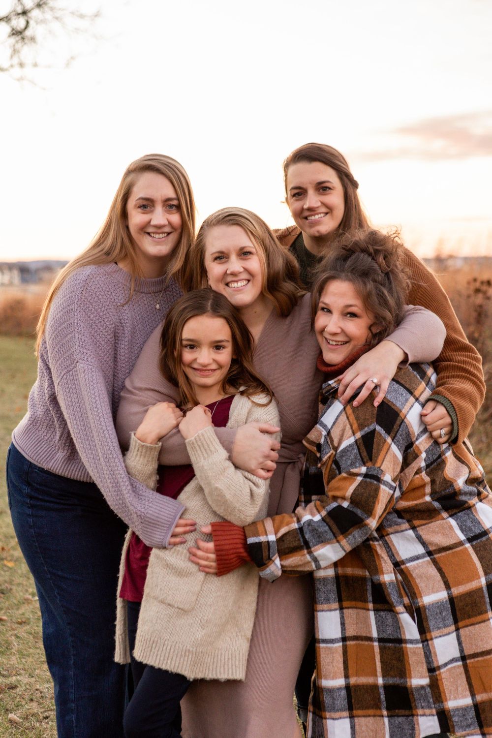 A group in warm sweaters and coats huddle together while embracing outdoors during sunset.