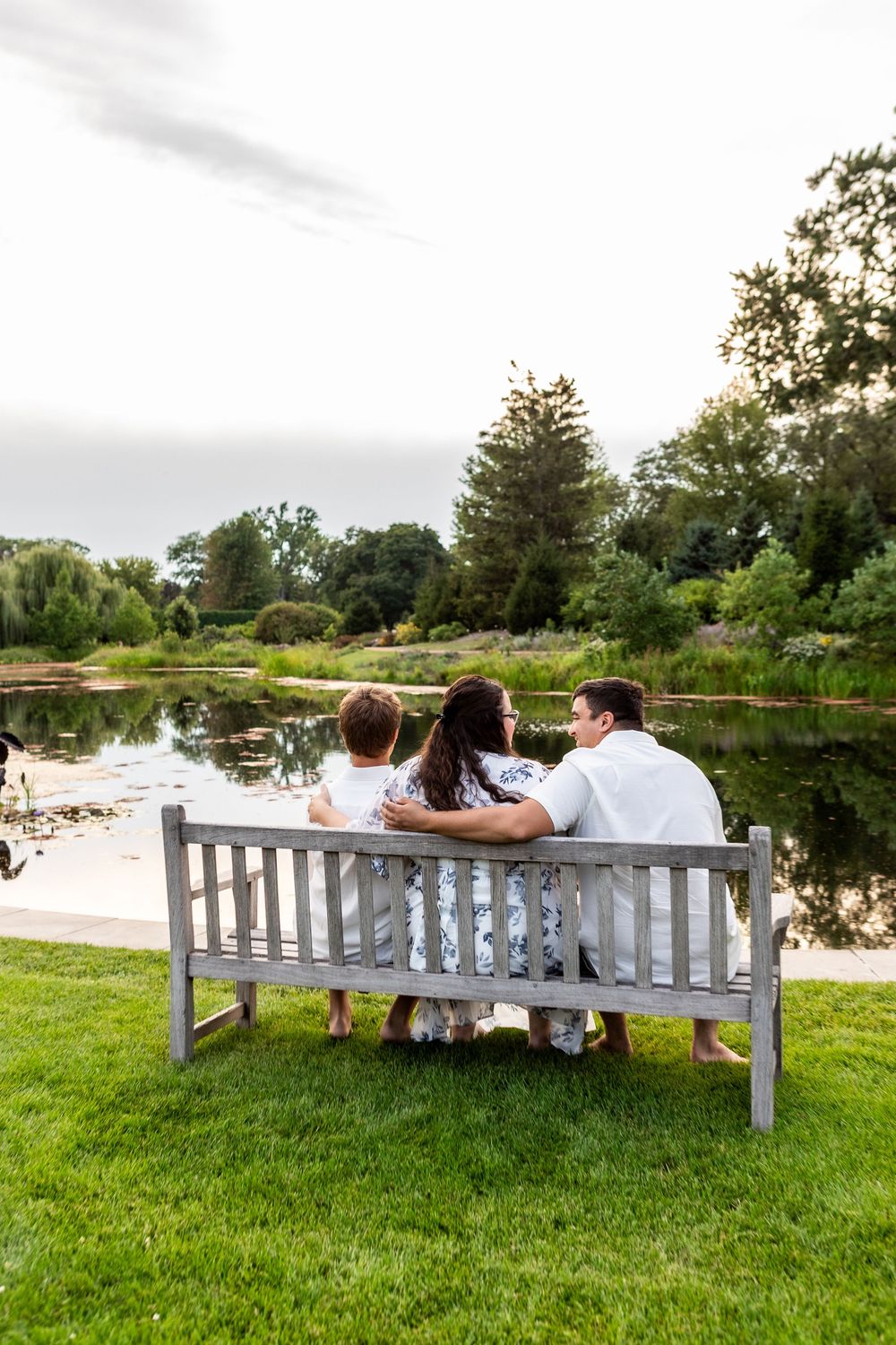 A family sits together on a wooden bench overlooking a serene pond surrounded by green grass and trees.