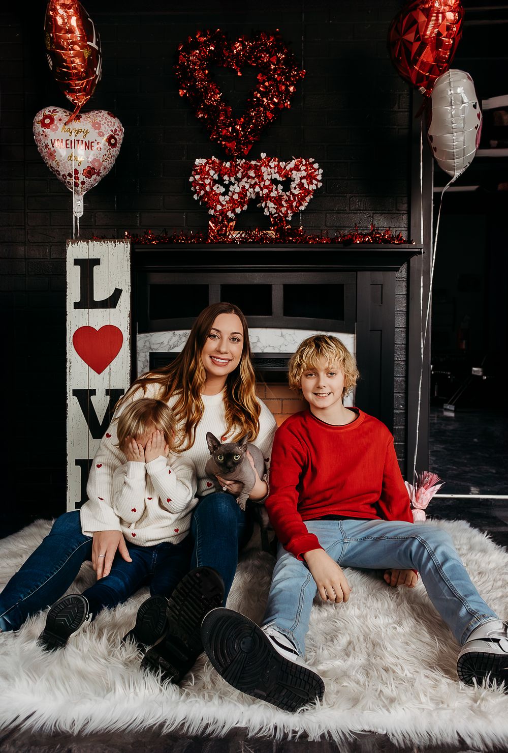 Woman and two children pose with a cat amid Valentine's Day decorations, balloons, and a LOVE sign on a fluffy rug.