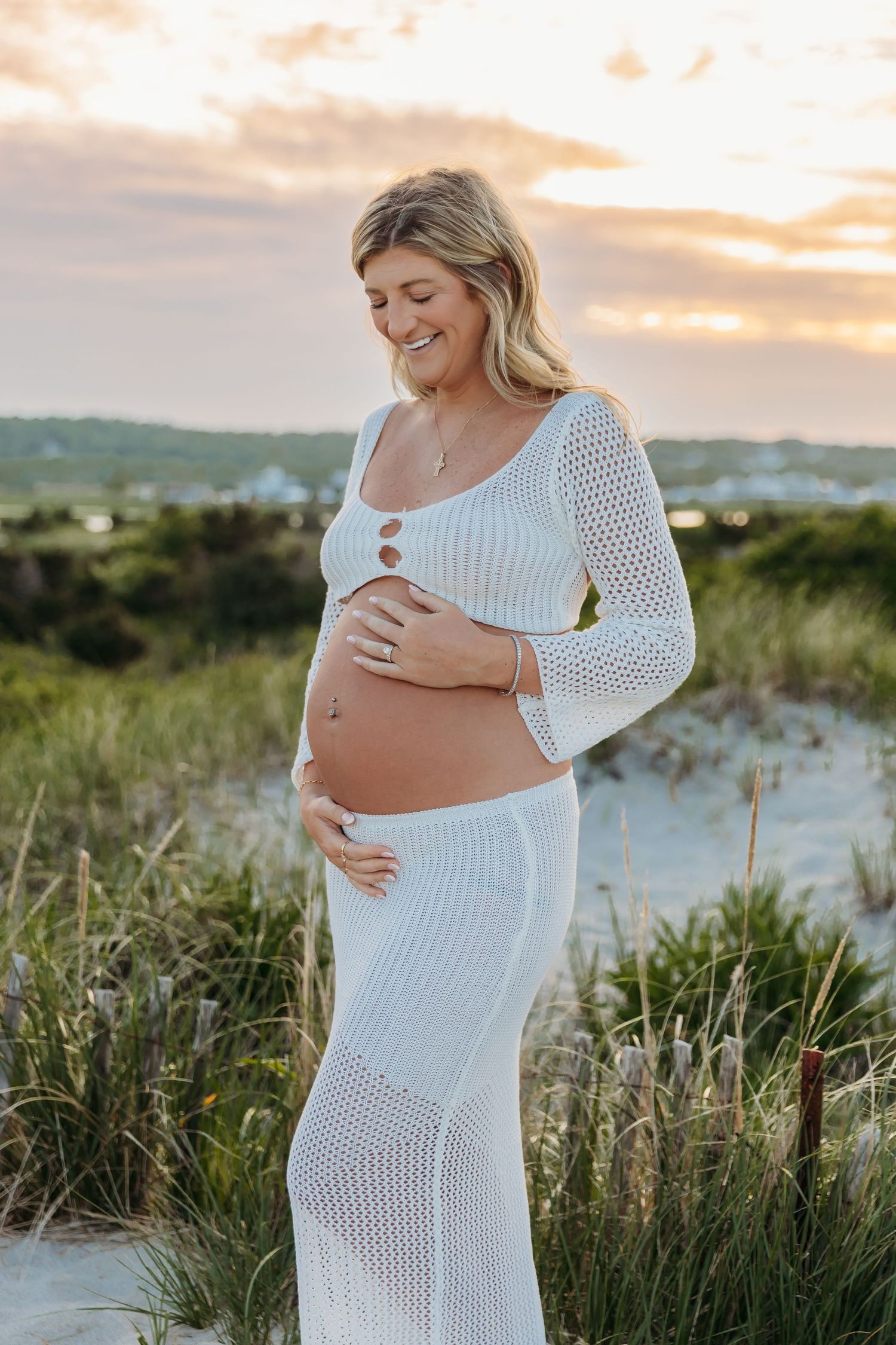 Expectant mother in white crochet dress poses on beach dunes at sunset while cradling baby bump.