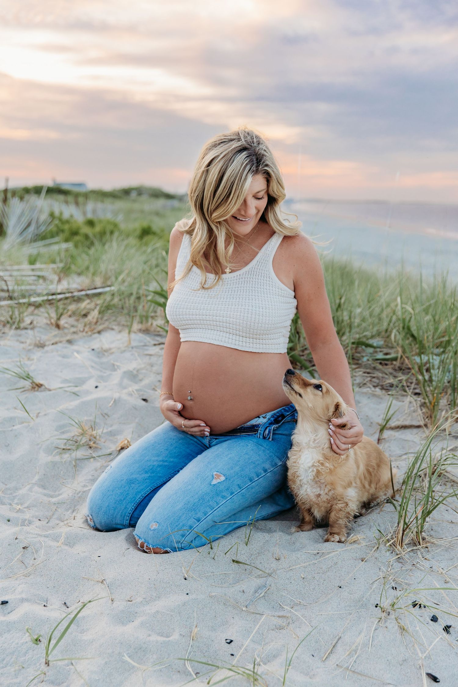 Expecting mother in white crop top and jeans sits with small dog on sandy beach at sunset with sea grass in background.