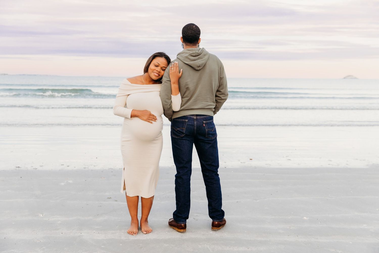 A couple stands together on a serene beach, the pregnant woman in a white dress against a cloudy coastal backdrop.