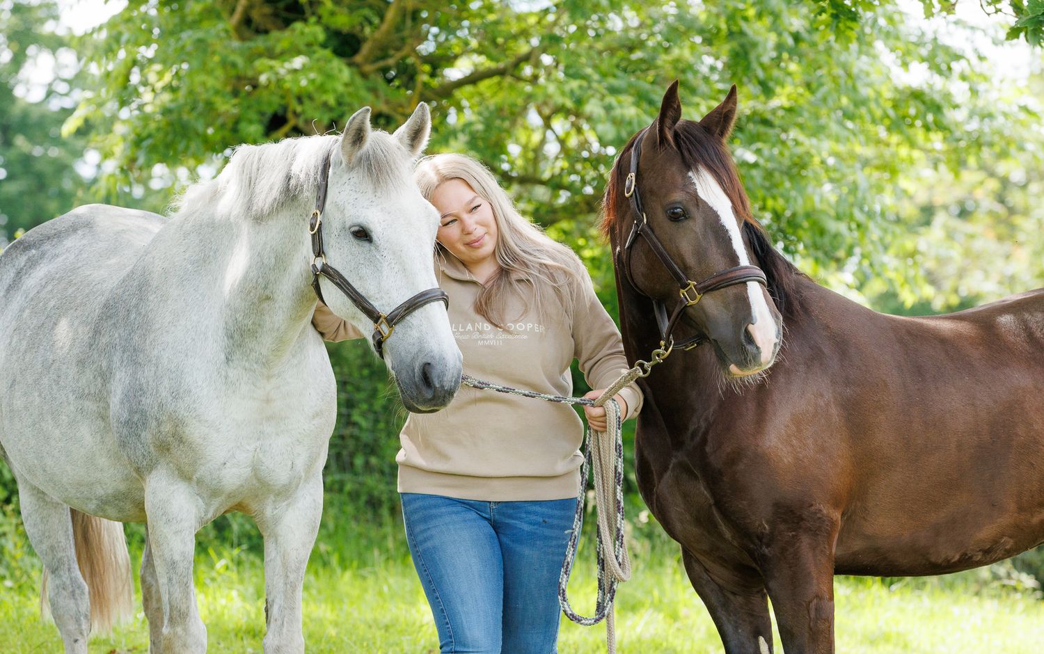 A woman sits on a log beside a brown horse in a field, holding its reins in a serene black and white photo.