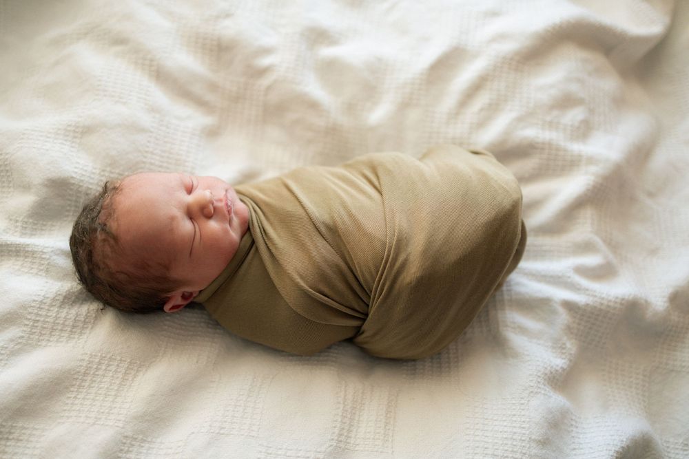 Newborn baby swaddled in olive green wrap sleeping peacefully on white textured blanket.