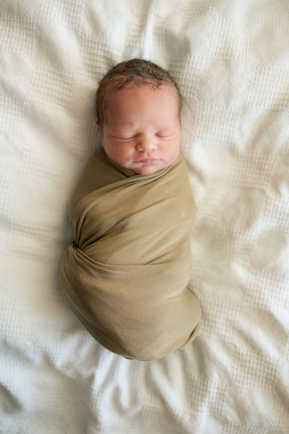 Newborn baby peacefully sleeping swaddled in tan wrap on white textured blanket.