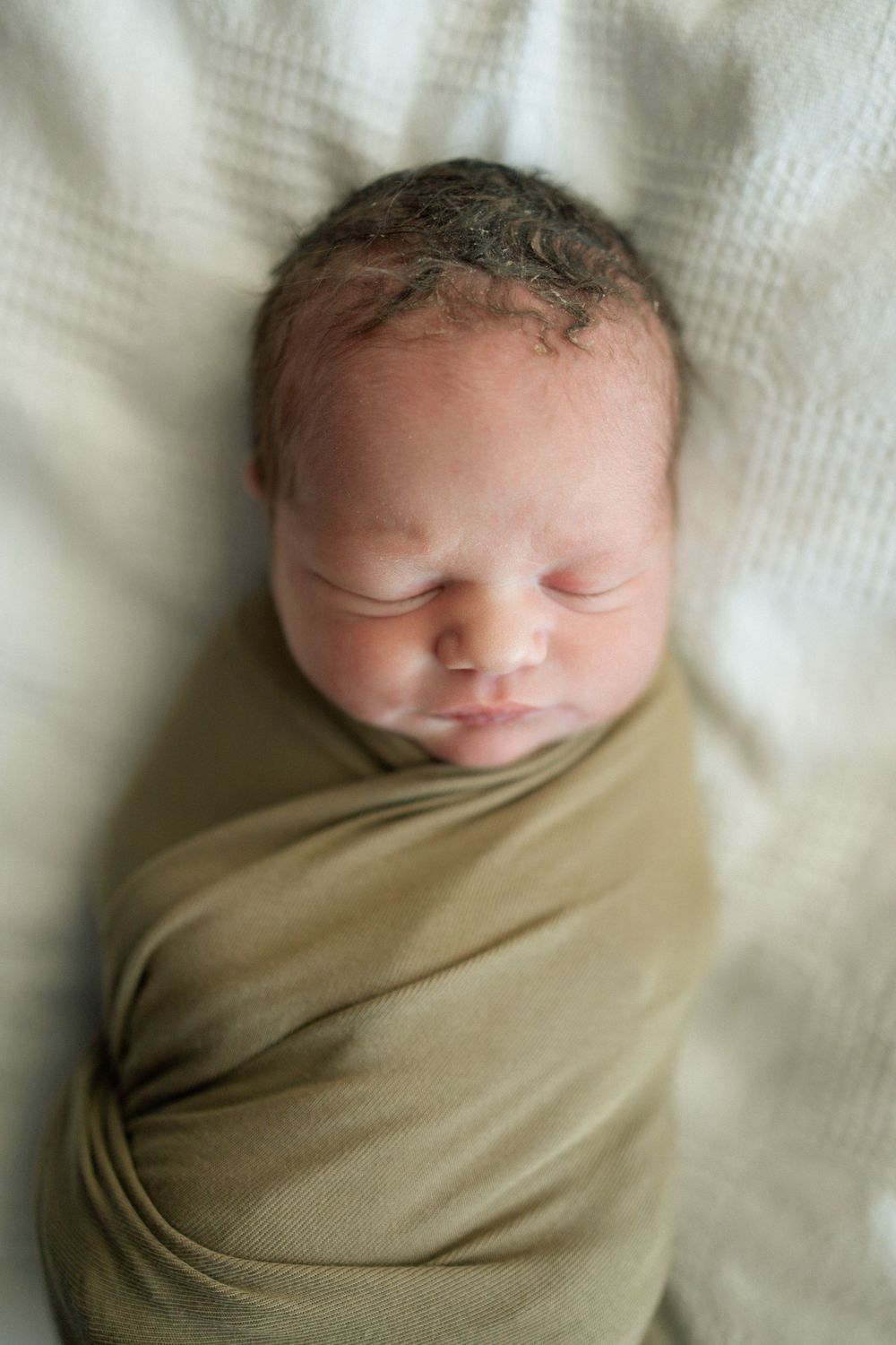 Peaceful newborn baby swaddled in olive green wrap sleeping on white textured blanket.