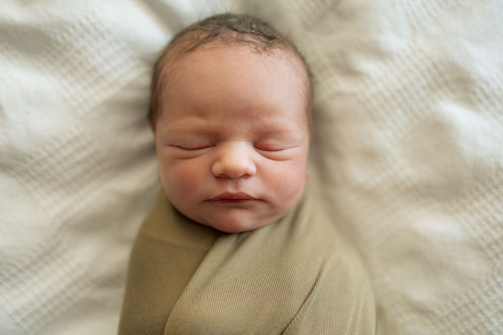 Newborn baby swaddled in a soft green wrap, sleeping peacefully on a white textured blanket.