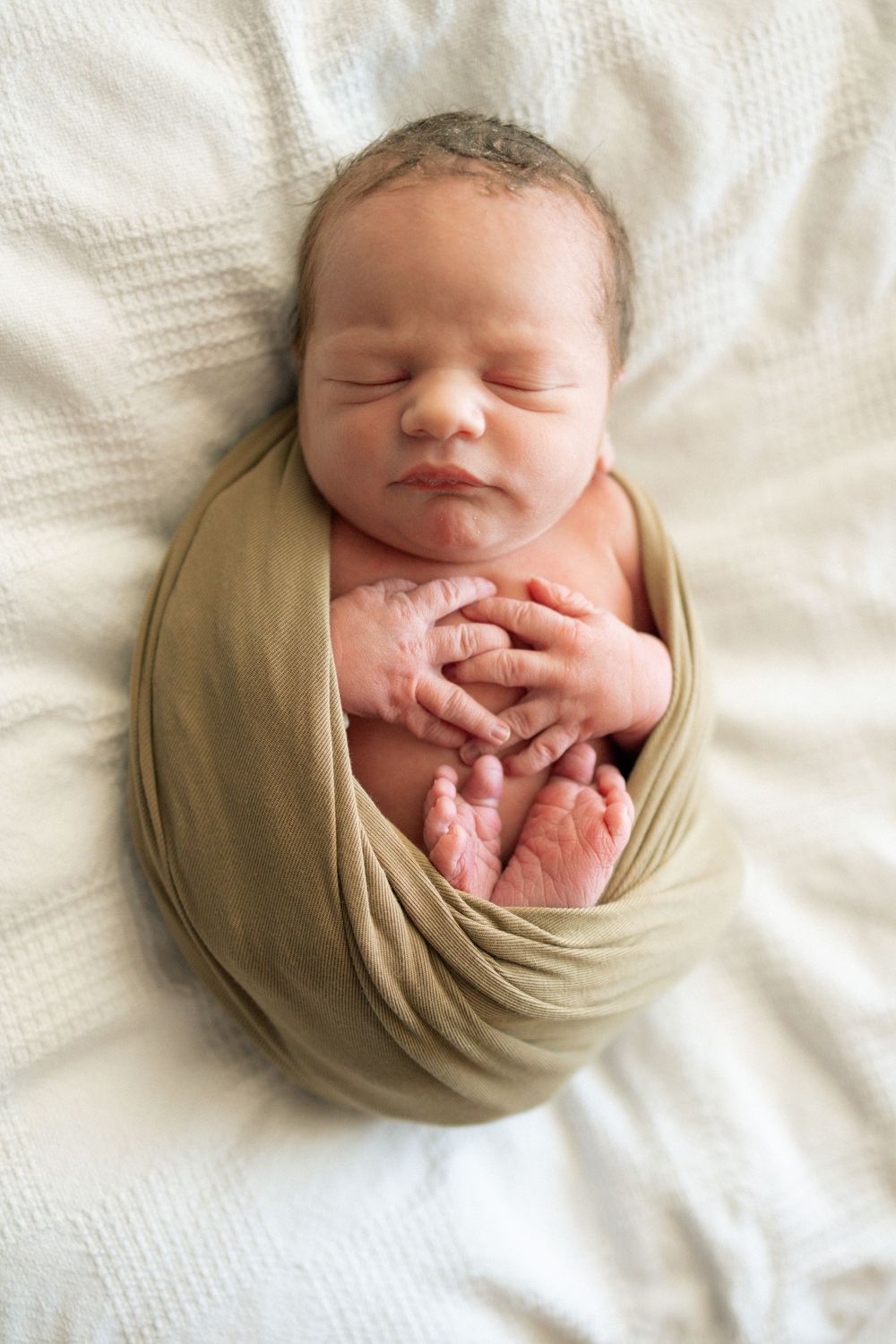 Sleeping newborn baby swaddled in olive green wrap with tiny hands and feet visible on white blanket.