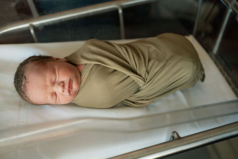 Newborn baby swaddled in olive green wrap sleeping peacefully in hospital bassinet.