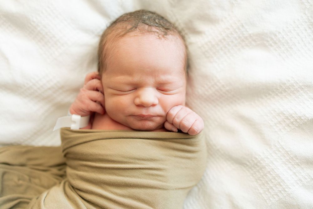 Peaceful newborn baby swaddled in tan wrap sleeping on white textured blanket.
