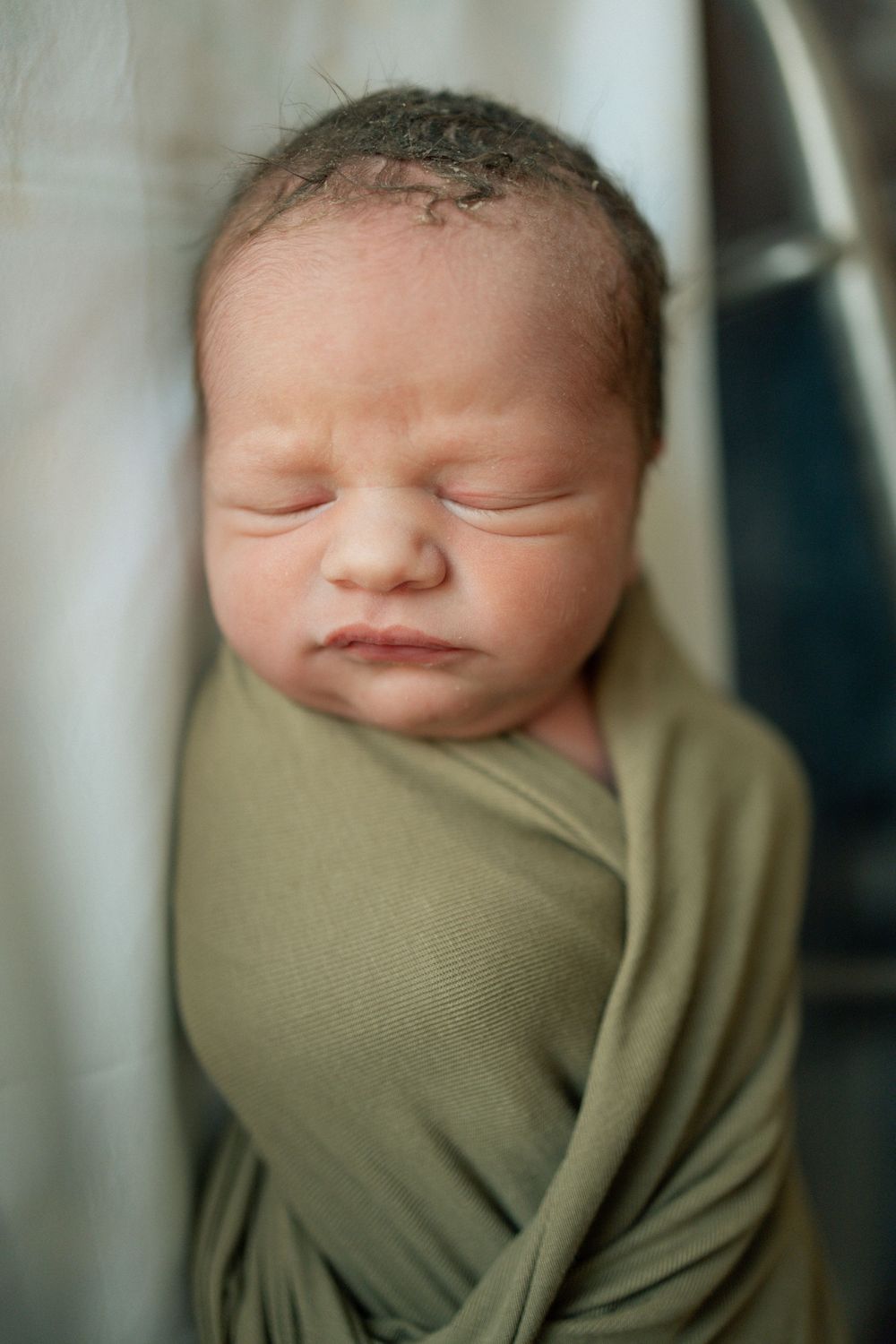 Sleeping newborn baby swaddled in olive green wrap with dark hair and peaceful expression.