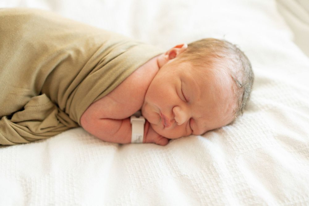 Peaceful newborn baby swaddled in olive green wrap sleeping on white blanket with hospital ID bracelet.
