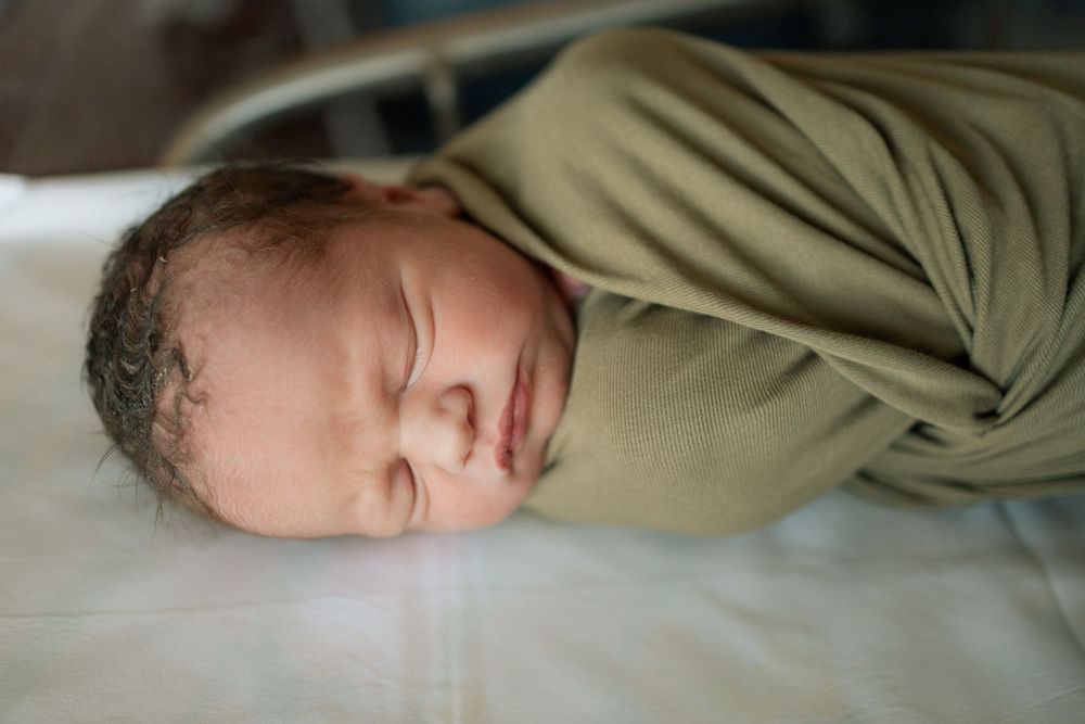 Newborn baby swaddled in olive green blanket sleeping peacefully on white surface in hospital.