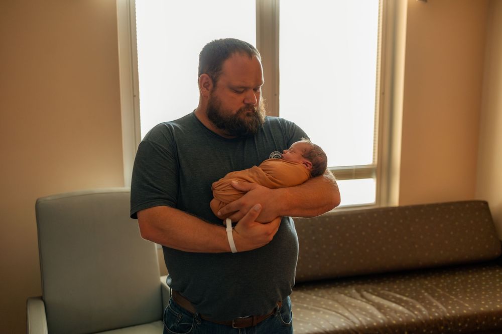 A bearded man in a gray shirt holds a swaddled newborn baby in a hospital room near a window.