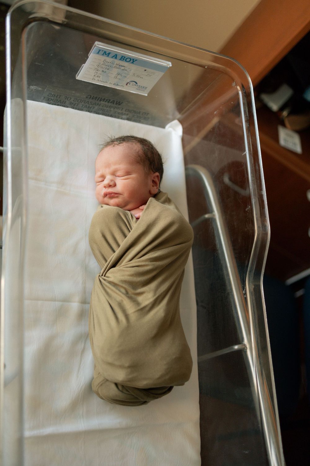 Newborn baby boy swaddled in olive green blanket sleeping peacefully in hospital bassinet with I'm a Boy label.