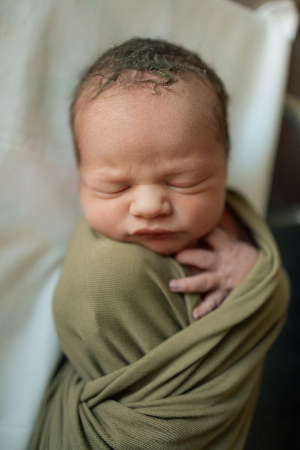 Sleeping newborn baby swaddled in olive green wrap with tiny hand visible against chest.