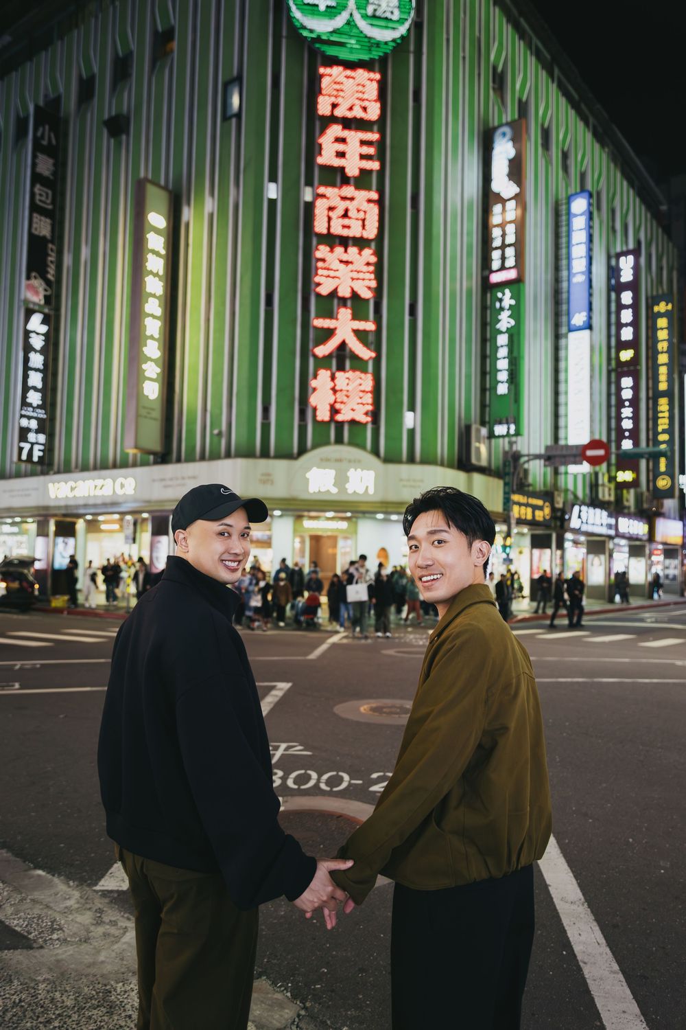 Two men holding hands on a city street at night in front of a neon-lit building with Chinese characters in Taiwan.