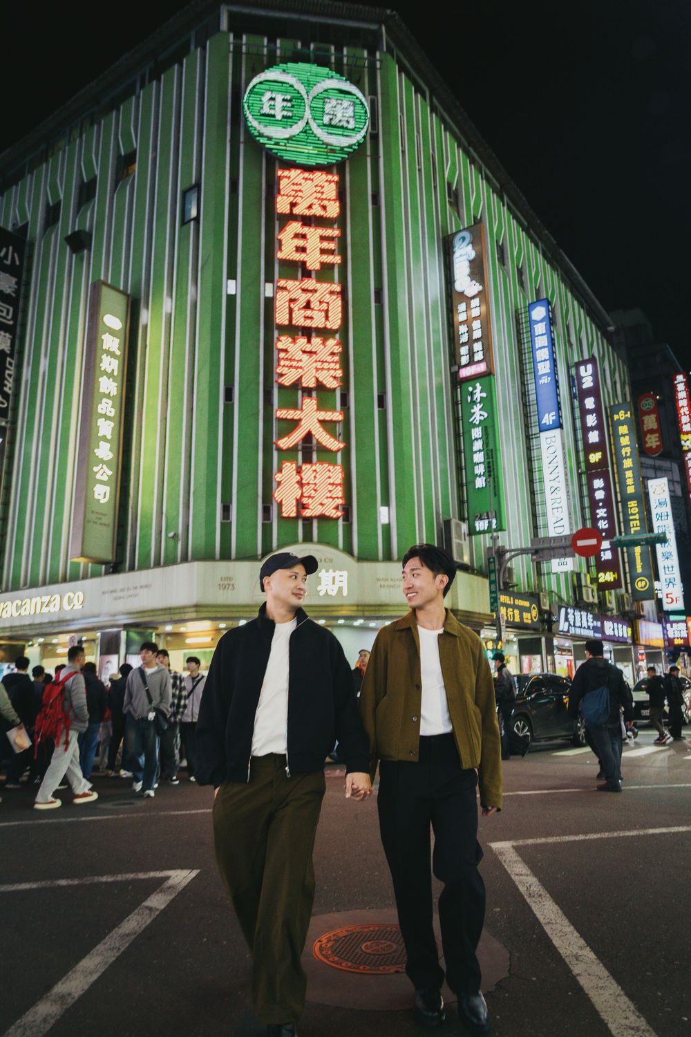 Two men walk hand in hand at night in a vibrant Taipei street with neon signs and a green-lit commercial building.