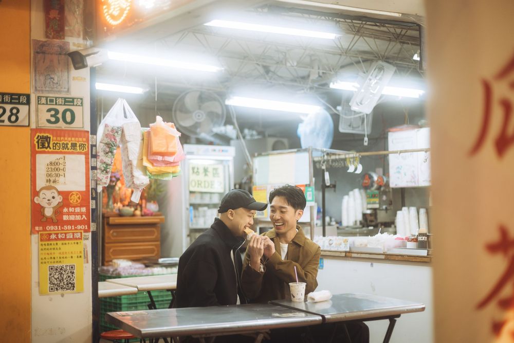 Two people eating at a traditional Asian street food stall with bright lights and Chinese signage.