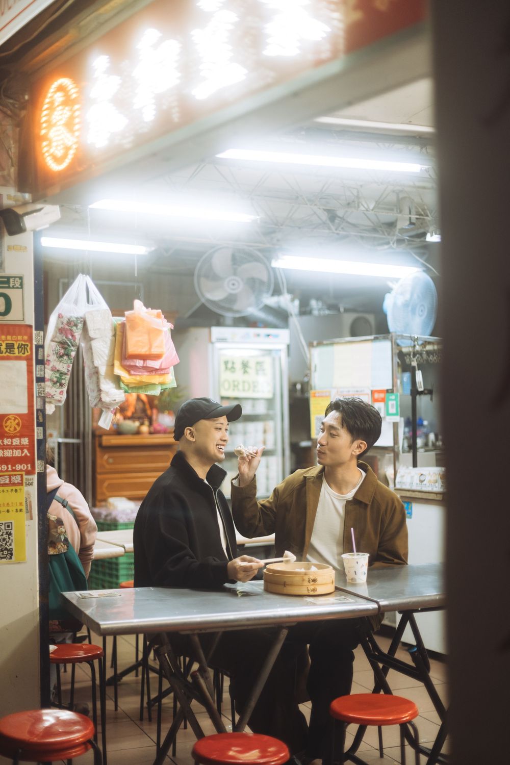 Two young men enjoying dim sum and conversation at a cozy Taiwanese street food stall at night.