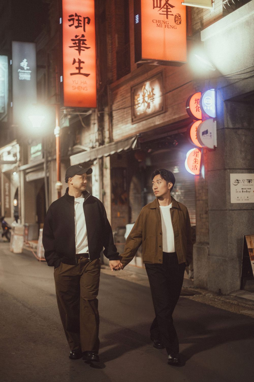 Two young men hold hands walking down a vibrant Asian city street at night, lit by neon signs.