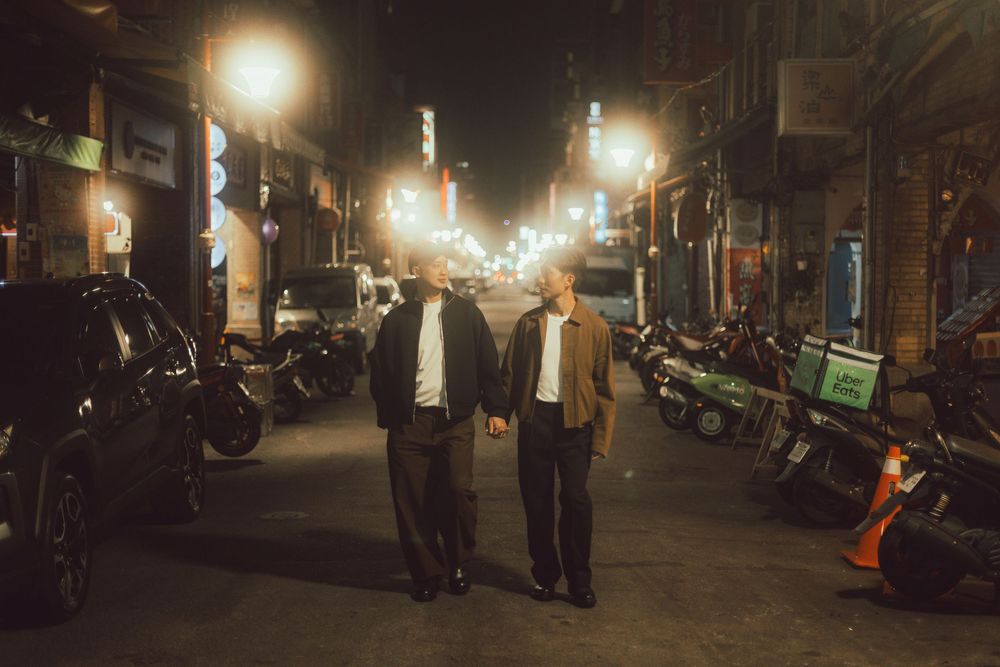 Two men walking together on a vibrant night street lined with motorcycles and glowing city lights.