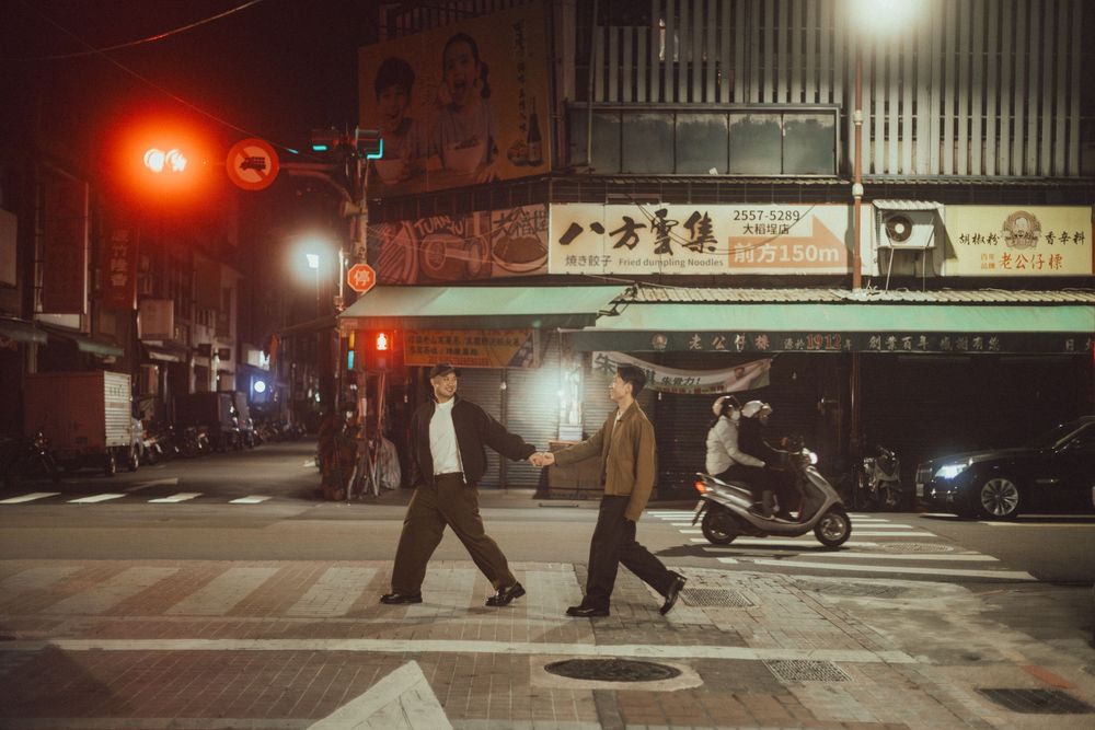 Two people crossing a street at night in an Asian city with red traffic lights and storefronts.