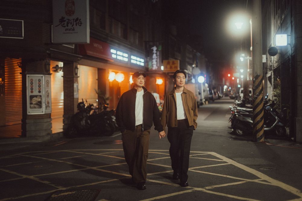 Two men walk hand in hand on a night street in Taiwan, lit by warm storefront lights and street lamps.