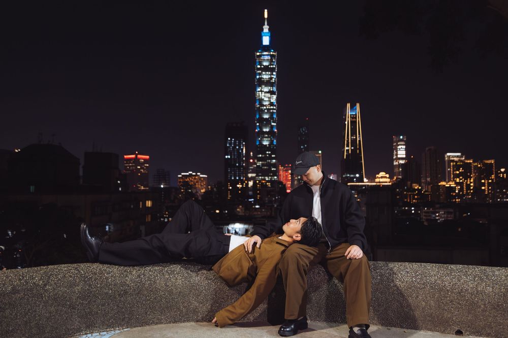 Two people relaxing on a stone ledge at night with Taipei 101 skyscraper illuminated in the city skyline background.