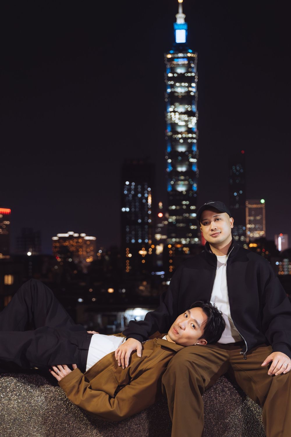 Couple relaxing on a rooftop at night with Taipei 101 tower illuminated in the background.