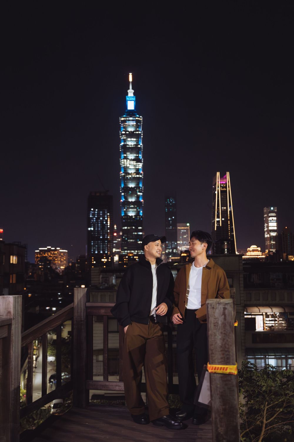 Two men stand on a rooftop terrace at night with Taipei 101 skyscraper illuminated in the background.