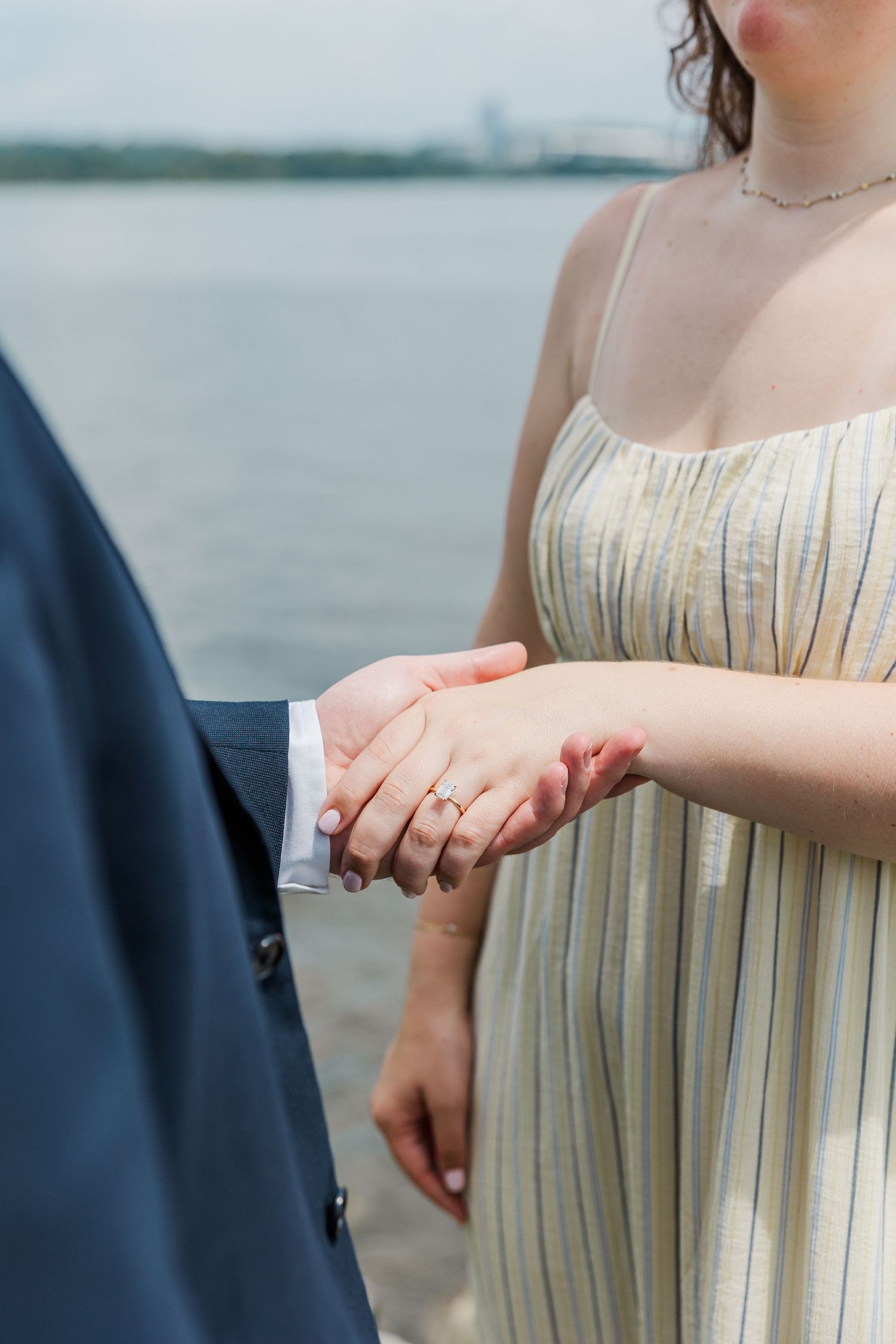 Couple holding hands near waterfront, woman wearing engagement ring and striped sundress, man in navy suit jacket.