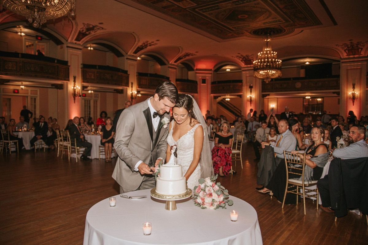 Bride and groom cutting wedding cake in elegant ballroom with warm lighting and guests seated at tables.