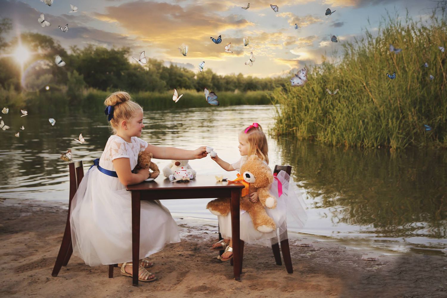 Two little girls share a tea party at a small wooden table on a sandy lakeshore at sunset with a teddy bear.