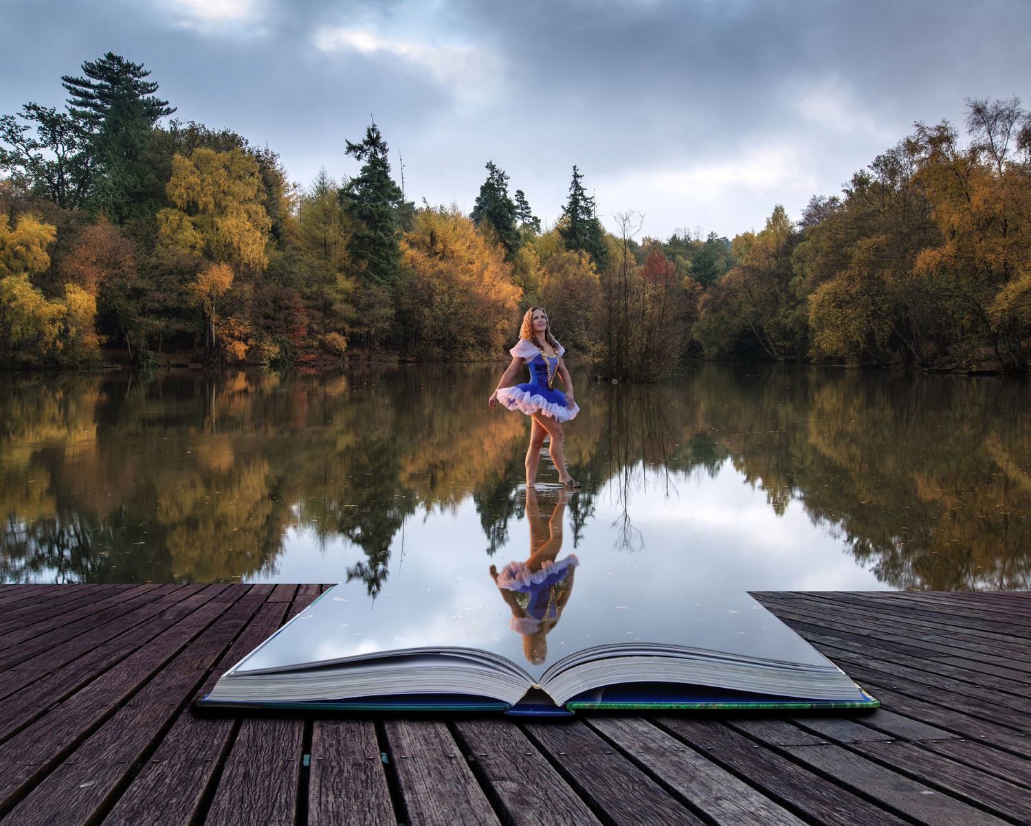 A ballerina in a blue tutu stands on an open giant book on a wooden dock overlooking a lake and autumn trees.