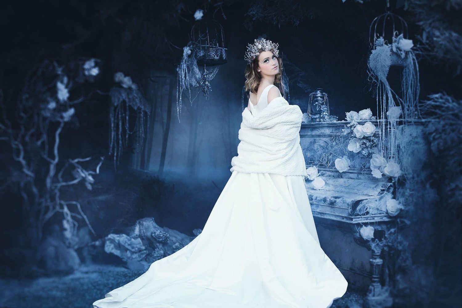 A woman in a flowing white gown and floral crown stands in a moody blue fantasy forest with hanging cages.