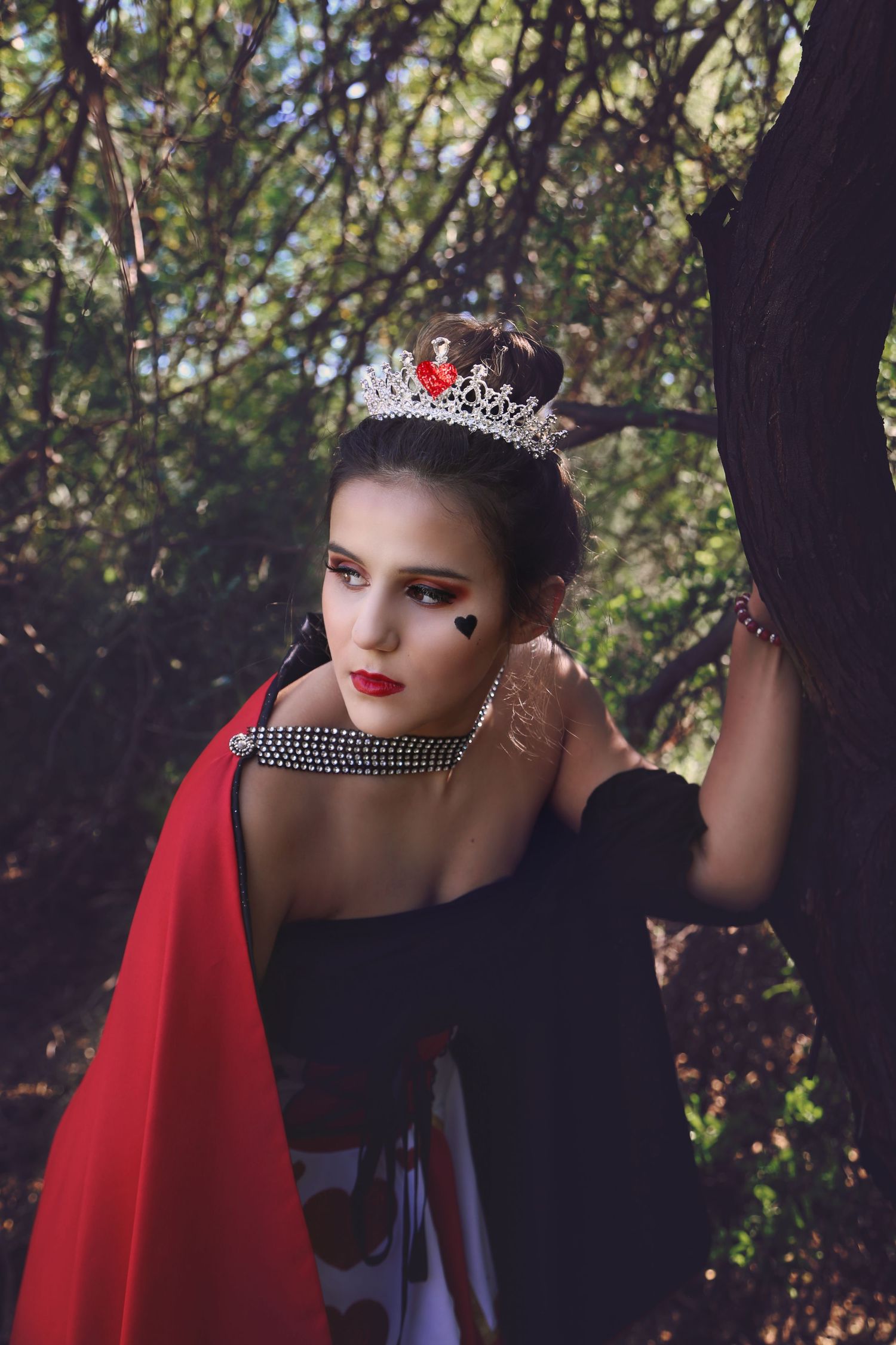 A woman dressed as the Queen of Hearts wears a sparkling tiara, red cape and heart face paint while leaning against a tree.