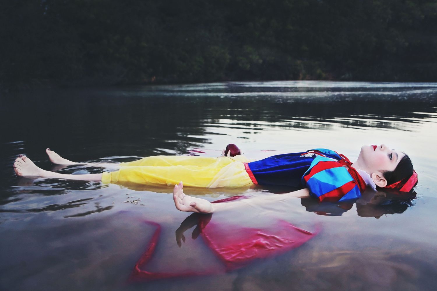 A woman dressed as Snow White floats on her back in dark lake water, her yellow skirt and red cape spreading around her.