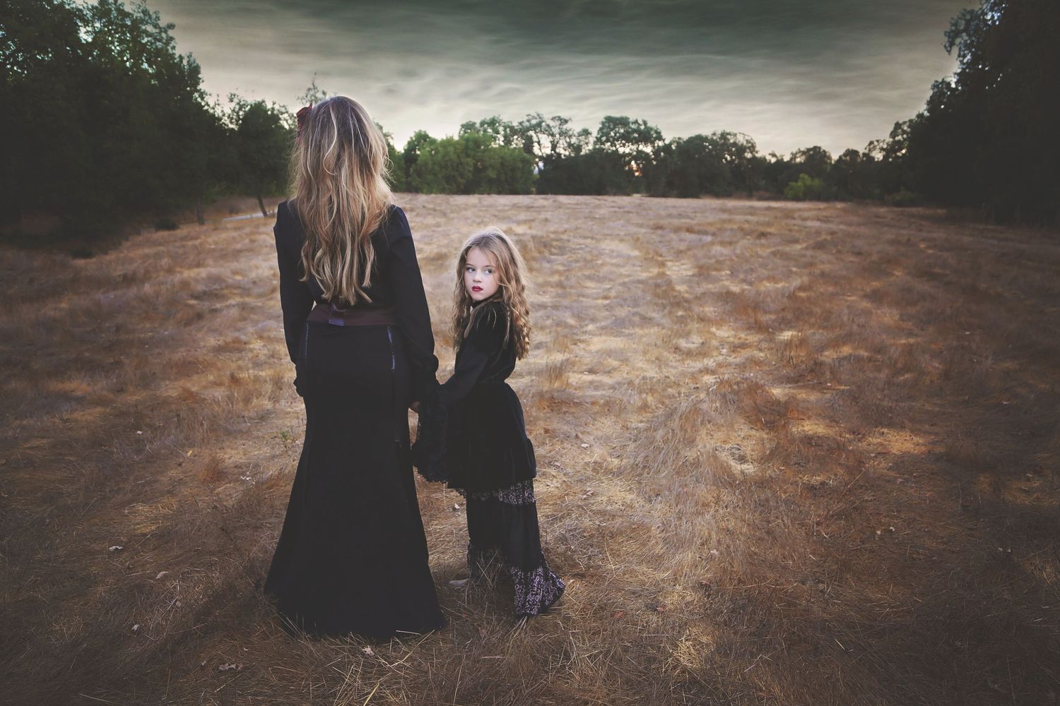 A woman and a young girl in matching black dresses hold hands and face a dry golden field under a moody, cloudy sky.